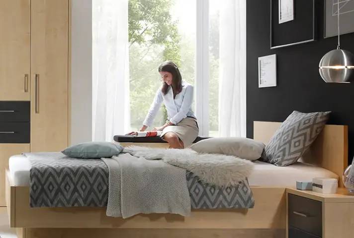 Schlafzimmer mit Holz-Bett und grauer Bettwäsche, Person sitzt vor hohem Fenster über offenem Koffer, links Kleiderschrank, rechts Nachttisch mit Lampe.